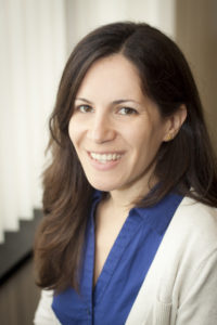 Erica is a White woman with brown hair. She is smiling and wearing a blue dress and cream cardigan. She is standing in front of an office window.