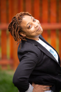 Acacia is a Black woman with medium-length locs and is standing in front of an orange fence. She is wearing a white shirt and dark blazer and is leaning back slightly to smile at the camera.