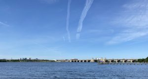 View of the Hanover Street Vietnam Veterans' Memorial Bridge from Middle Branch Park. The water has gentle ripples and the sky is bright blue with clouds. A small, wooden pier and the Baltimore skyline are also in the distance.