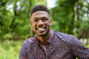 Ethan is a Black man with medium length dark brown hair. He is smiling, wearing a dark blue shirt with a floral pattern, and is standing in front of the forests of Druid Hill Park in Baltimore, MD.