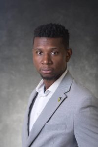 Justin is a Black man with a dark brown complexion and medium length hair. He is wearing a gray suit jacket, white collared shirt, and lapel pin. He is standing in front of a gray backdrop and is looking directly at the camera.