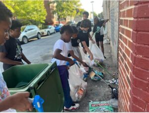 Children with trash grabbers and white garbage bags are cleaning up litter along a sidewalk next to rowhouses. This group is part of My Father's Plan's neighborhood clean-up program, supported by SBGP's Enhanced Services' Waste Management funding.
