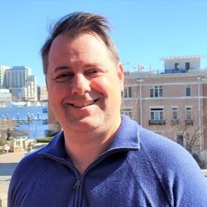 Jeff is a white man with short light brown hair. He is smiling and wearing a blue collared sweater with a zip. He is standing on a deck in front of reddish brick buildings and the Baltimore harbor, skyline, and bright blue sky in the background.