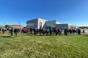View of the grand opening of the Middle Branch Fitness and Wellness Center. People are gathered outside the gray, blocky building on the grass and the sky is bright blue.