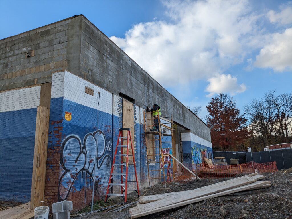Exterior view of the Carroll Park Rec Center renovation. The graffiti-covered building has ladders and scaffolding next to it with a construction worker working on the roof, which is being removed.