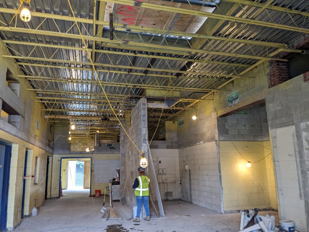 View of the inside of the Rec Center which has been completed gutted. The walls are bare cinder blocks and the shiny, ceiling paneling is visible between yellow support bars. Construction workers are visible in the bright yellow vest and hard hats under strings of work lights.