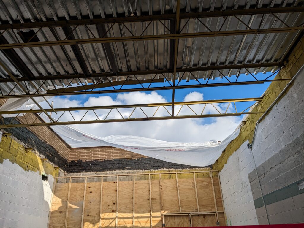 A view of the blue sky is visible through the open roof of the Rec Center that is being replaced. The interior demolition shows exposed I-beams on the ceiling and wooden studs and plywood of the walls.
