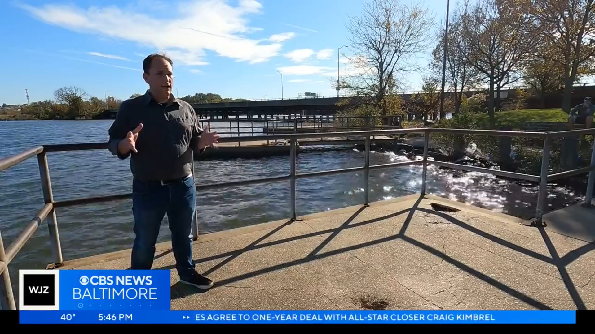 Screenshot from the interview of Brad Rogers, a white man wearing a gray shirt and jeans, standing on a pier in the Middle Branch with the waterfront and bridge in the background.
