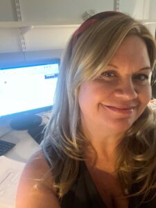 Jennifer is a white woman with blond hair and a red and black checkered headband. She is smiling in front of a blurred computer screen in an office.