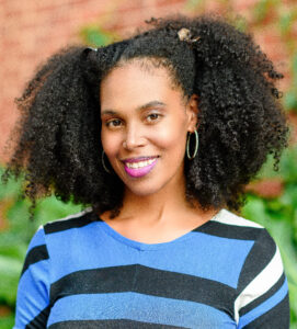 Kanika is an African American woman with medium long dark hair, styled in a curly afro that frames her face. She is smiling and wearing a blue, black and white bold-striped dress, purple lipstick, and silver hoop earrings. She is standing in front of a brown brick wall and green foliage.