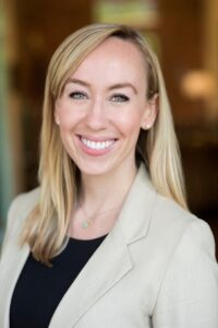 Samantha is a white woman with blonde hair, green eyes, and a big smile. She is wearing a tan blazer over a black top, accessorized with pearl earrings and a delicate, seafoam green necklace. She stands in front of an artistically blurred background.