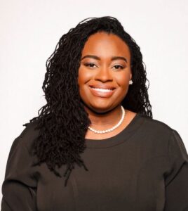 Ashley is a black woman with dark dreaded hair and white pearls around her neck. She is smiling and wearing a black dress and pearl earrings. She is sitting in front of a white background.