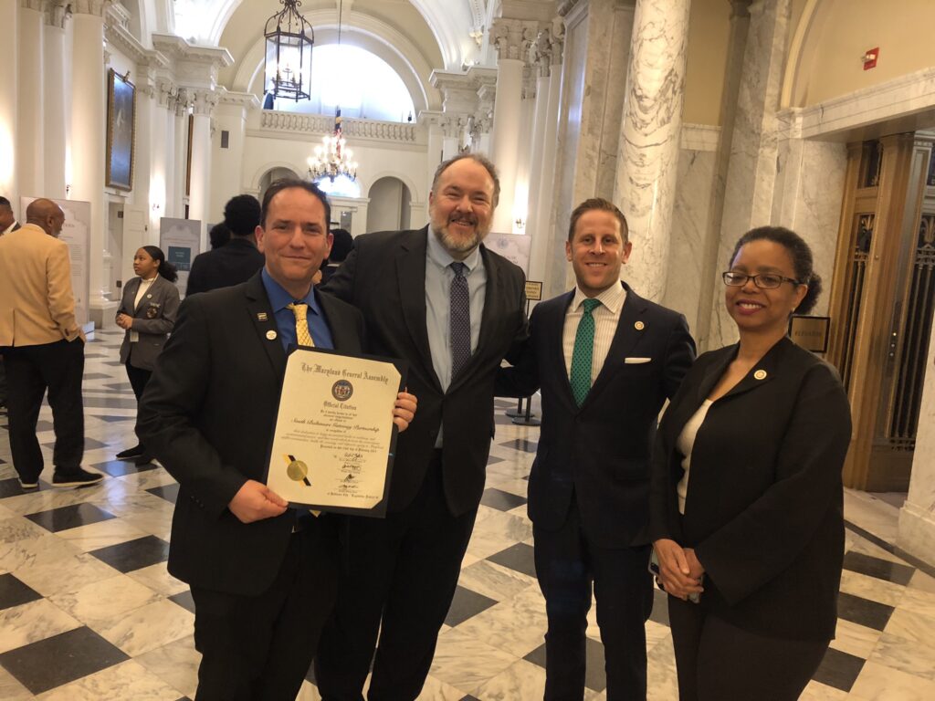 Brad Rogers from SBGP (holding the official citation, Delegate Clippinger, Delegate Edelson, and Delegate Lewis are in the House hallway smiling at the camera.