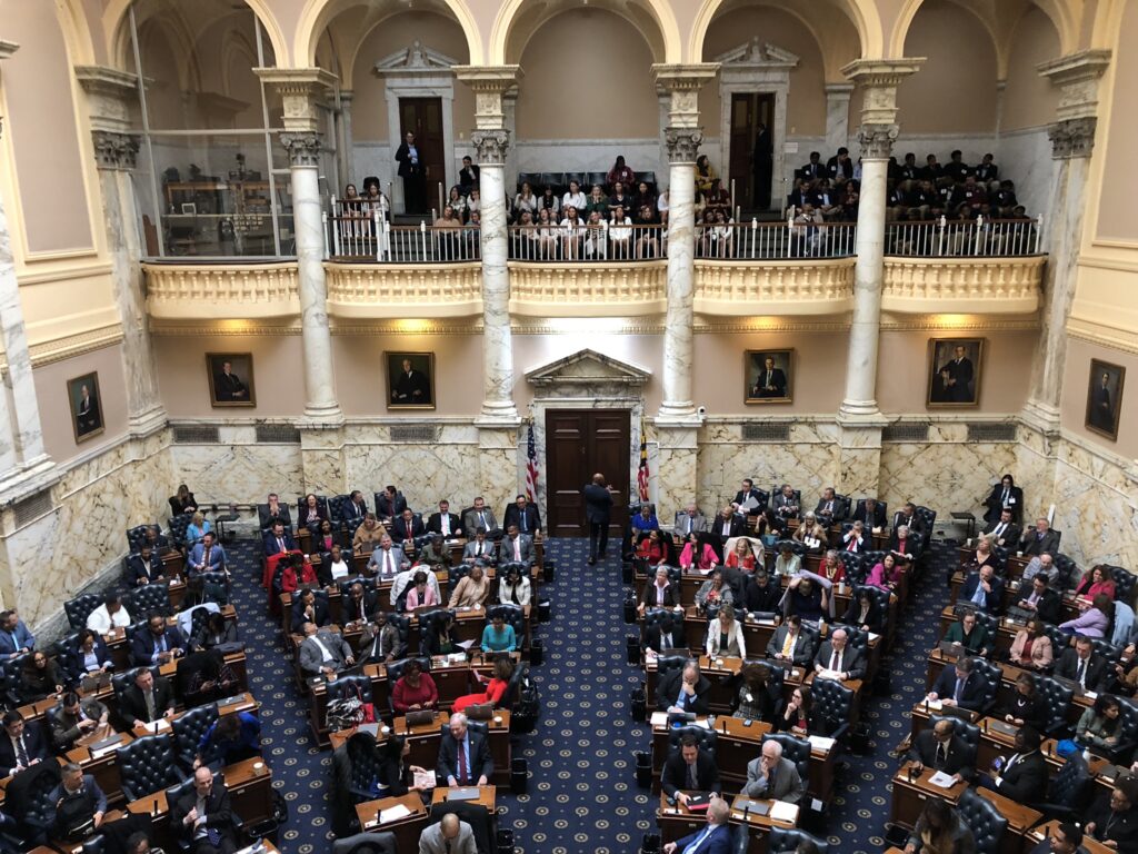 View of the Maryland Delegation Floor from the balcony of the House. The whites walls are lined with columns and people are visible sitting on the ground floor and across on the opposite balcony.