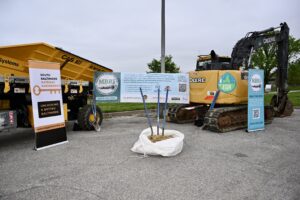 View of the Middle Branch Resiliency Initiative banner between large, yellow construction equipment, next to an SBGP banner and a larger bag of sand with shovels stuck in it.