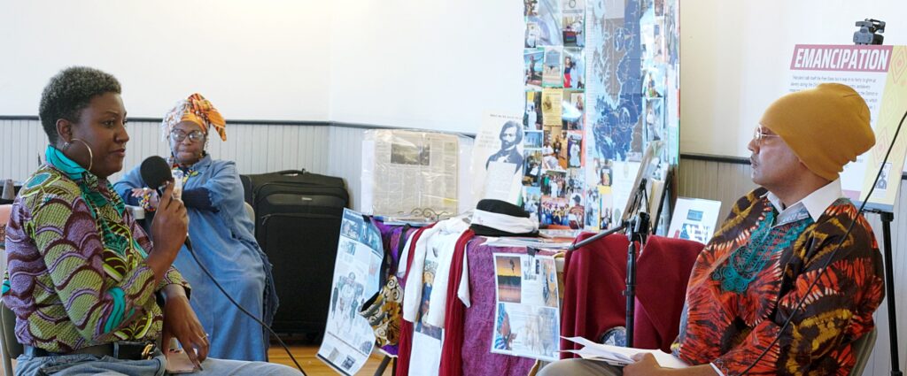 Panelist Asantewa Boakyewa is wearing a colorful shirt and gold hoop earrings, sitting with a microphone during the panel across from Fanon Hill. Fanon is also seated, wearing a colorful shirt and yellow hat.