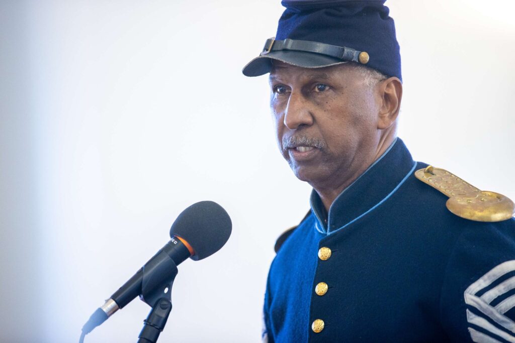 A re-enactor portraying a soldier from the United States Colored Troops (USCT) is speaking at a microphone wearing a navy blue cap and uniform with brass buttons.