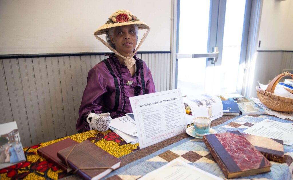 Female RE-Enactors of Distinction (FREED) re-enactor Lavonda Broadnax is sitting in a chair at an information table about Frances Ellen Harper Watkins. She is wearing a beautiful, period-appropriate outfit consisting of a purple dress and elaborate bonnet.