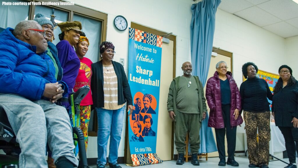 A group of community residents and partners are gathered and smiling next to one of the new banners, including Ms. Betty Bland-Thomas, Community President for Historic Sharp-Leadenhall.